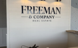 Custom dimensional Freeman & Company Real Estate lobby logo installed on a white shiplap feature wall with accent lighting on Long Beach Island, New Jersey